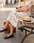 A woman sitting on a wooden park bench wearing a cream floral midi dress, black Mary Jane flats, and a straw boater hat with a black band. A brown leather handbag sits beside her on a paved outdoor walkway.
