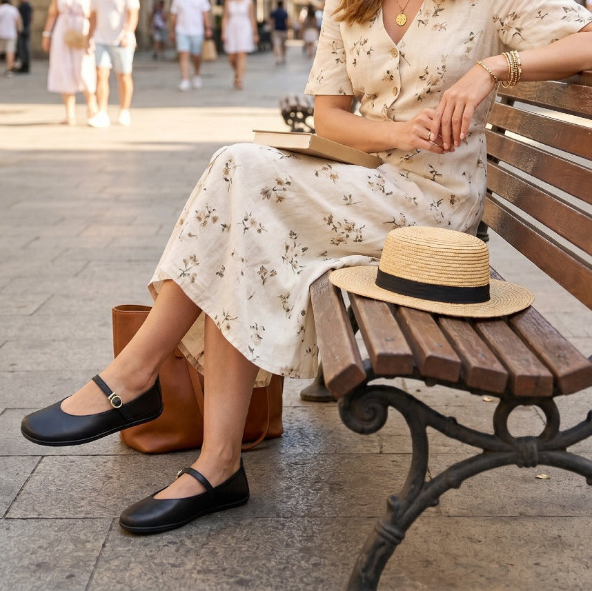 A woman sitting on a wooden park bench wearing a cream floral midi dress, black Mary Jane flats, and a straw boater hat with a black band. A brown leather handbag sits beside her on a paved outdoor walkway.