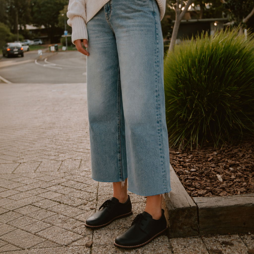 A person standing outdoors on a paved sidewalk wearing light-wash cropped wide-leg denim jeans, a cream knit sweater, and black lace-up shoes.