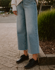 A person standing outdoors on a paved sidewalk wearing light-wash cropped wide-leg denim jeans, a cream knit sweater, and black lace-up shoes.